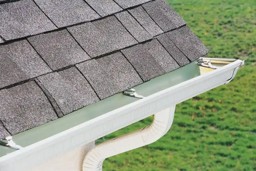 White seamless aluminum gutter and downspout outlet along a shingled roof edge