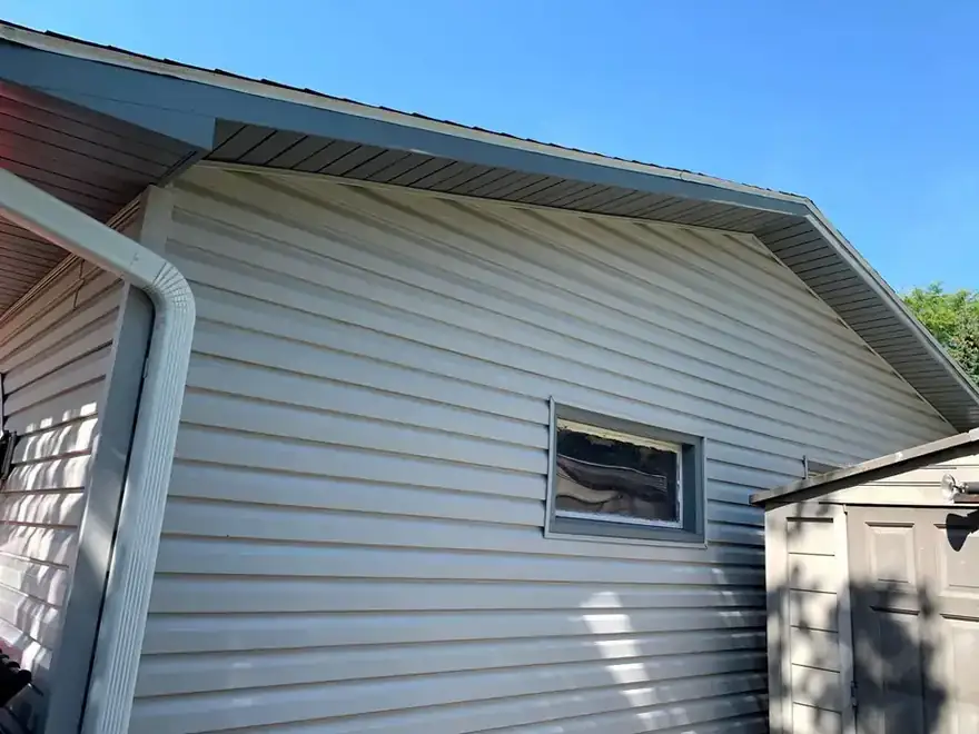 Gray vinyl siding under the eave with a white downspout against blue sky
